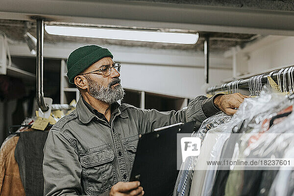 Bearded male tailor with clipboard checking clothes hanging on rack in workshop