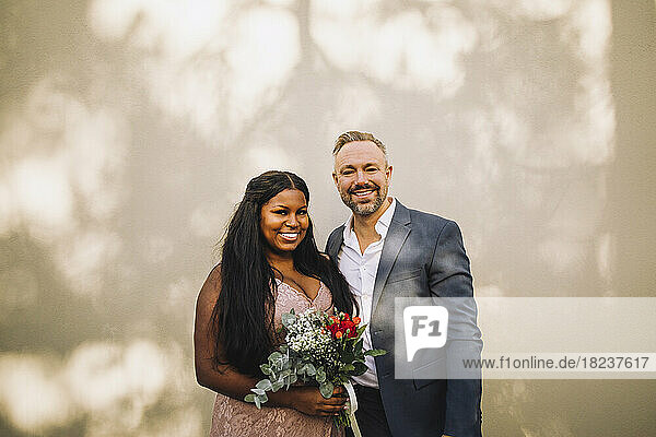 Portrait of smiling bride holding bouquet standing by groom against wall