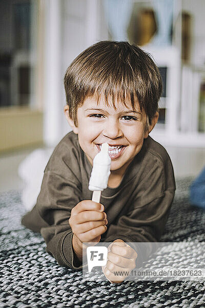 Porträt eines lächelnden Jungen  der auf einem Teppich liegend ein Eis isst