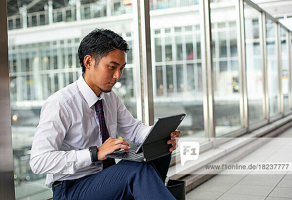 A young businessman in the city  on the move  sitting on a walkway  using his laptop.