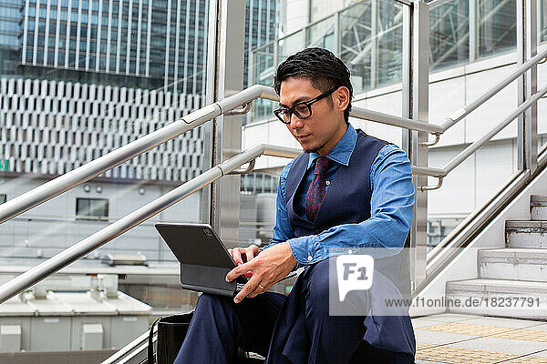 A young businessman in the city  on the move  seated on the stairs by a window  using a laptop.