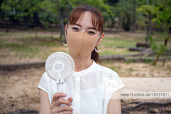 A mature Japanese woman outdoors in a park  wearing a face mask holding a small electric fan.