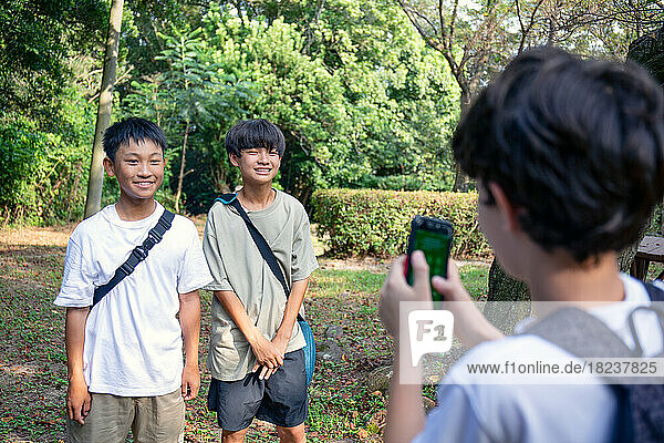 A boy with a mobile phone taking a picture of two 13 year old boys side by side  outdoors in a park in summer.
