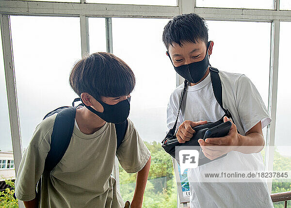 Two 13 year old Japanese boys in face masks  friends looking at a mobile phone screen.