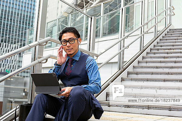 A young businessman in the city  on the move  sitting on stairs by a window on the phone  laptop open.