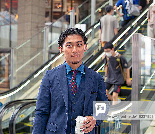 A young businessman in a blue suit on the move in a city downtown area  carrying a briefcase and cup of coffee.