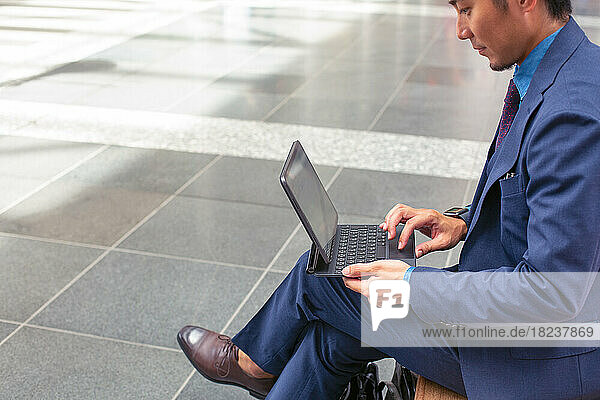 A young businessman in a blue suit on the move in a city downtown area  sitting on a bench using a digital tablet with a screen.