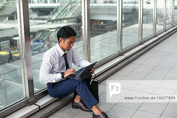 A young businessman in the city  on the move  sitting on a walkway  using his laptop.