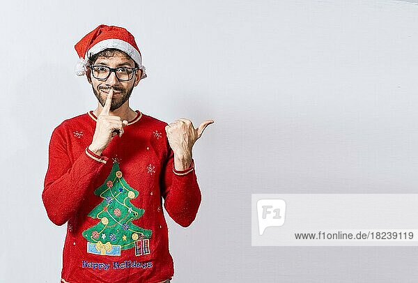 Mann in Weihnachtspullover in Stille Geste zeigt auf die Seite  Guy in Weihnachtsmütze mit Finger auf den Lippen zeigt auf die Seite. Bärtiger Mann in Weihnachtsmütze in der Stille Geste zeigt Promo