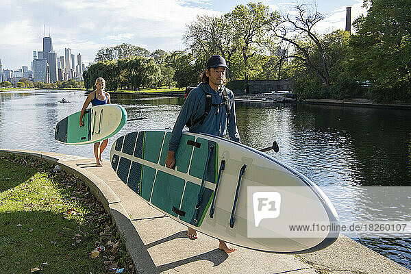 Man and woman carrying paddleboards walking at riverbank on sunny day