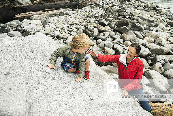 A mom helps her young son climb a boulder at a rocky beach