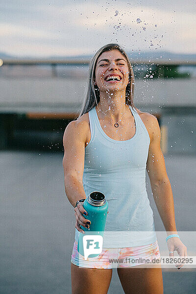 Fit woman splashing drink out of water bottle outside while smil