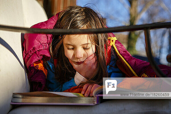 A little girl lays on couch outside in sleeping bag reading