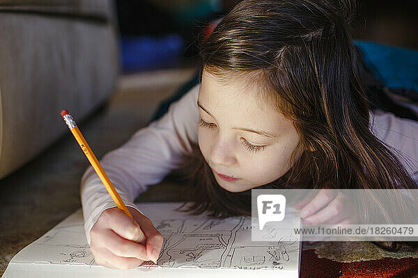 A little girl lays on floor carefully drawing in a sketchpad