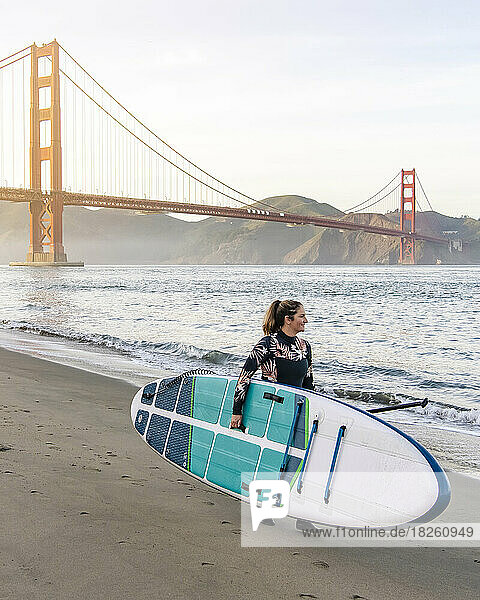 Woman carrying paddleboard walking towards Golden Gate Bridge