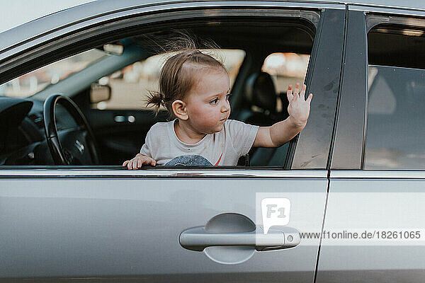 Toddler girl leaning out car window waving goodbye