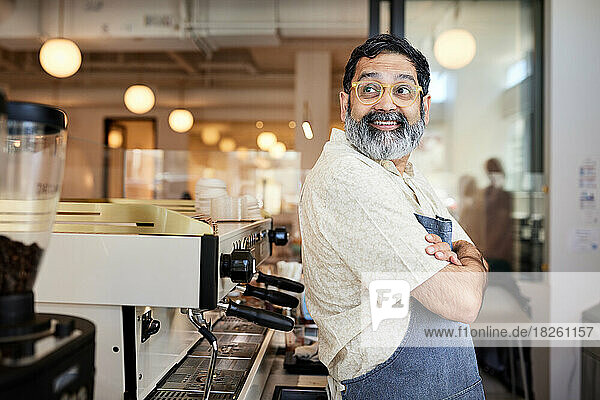 Smiling male barista wearing eyeglasses leaning on coffee machine