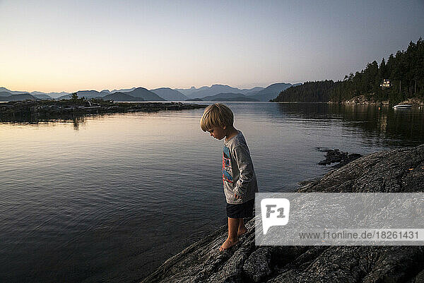 A young boy stands on a rock at water's edge at dusk