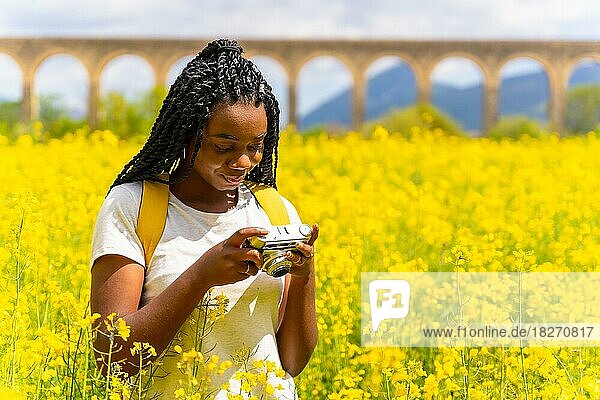 Looking at the photos on a vintage camera  a black ethnic girl with braids  a traveler  in a field of yellow flowers