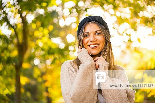 Junges blondes Mädchen mit einer Wollmütze im Herbst bei Sonnenuntergang in einem Naturpark lächelnd