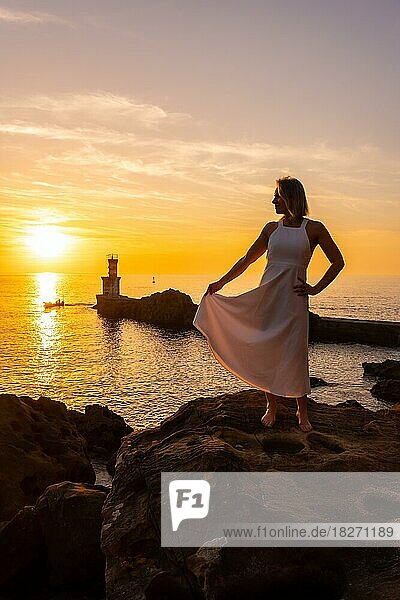 A blonde woman in a white dress at sunset next to a lighthouse in the sea  healthy and naturist life  lifestyle