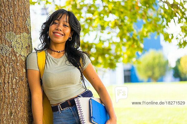 Asiatisches Mädchen auf dem Campus  lächelnd an der Universität unter einem Baum mit Block in der Hand und Rucksack