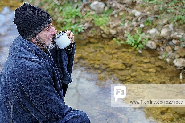 Bärtiger  mit einer Decke bedeckter Mann trinkt Kaffee am Flussufer
