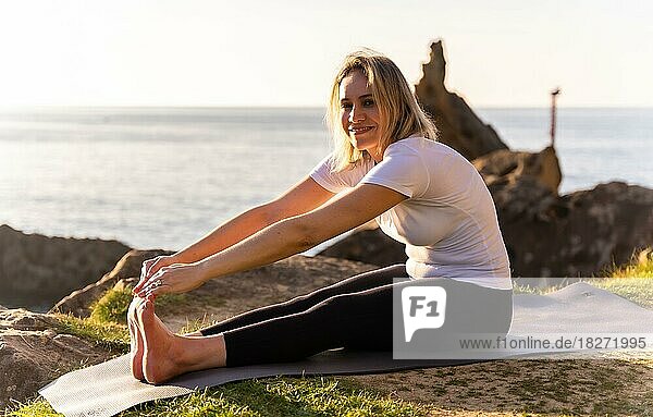 Eine blonde Frau macht Yoga-Übungen in der Natur am Meer  Stretching neben einem Leuchtturm  gesundes und naturistisches Leben  Pilates im Freien