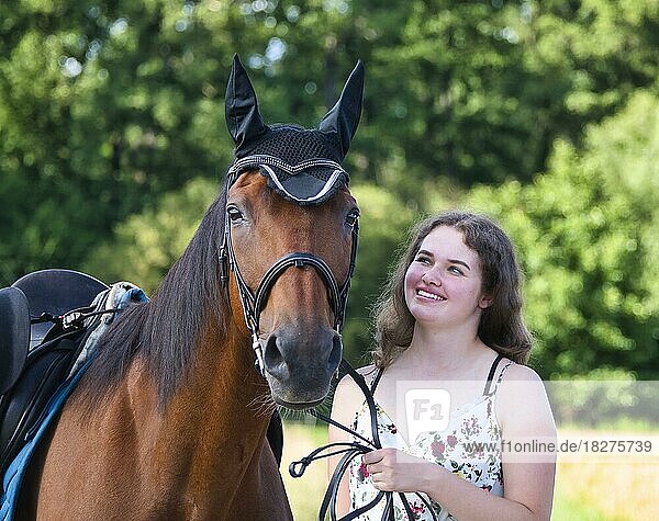 Portrait eines Mädchens im Teenageralter in einem Kleid  sie hält ein braunes Pferd am Zügel und schaut lächelnd zu ihm  das Pferd hat Sattel und Trense