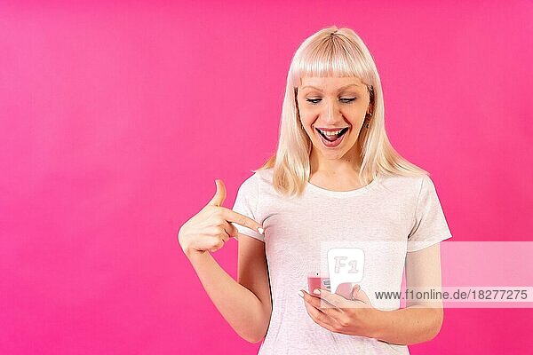 Blondes kaukasisches Mädchen im Studio auf rosa Hintergrund  Kopierraum  lächelnd auf das Handy zeigend