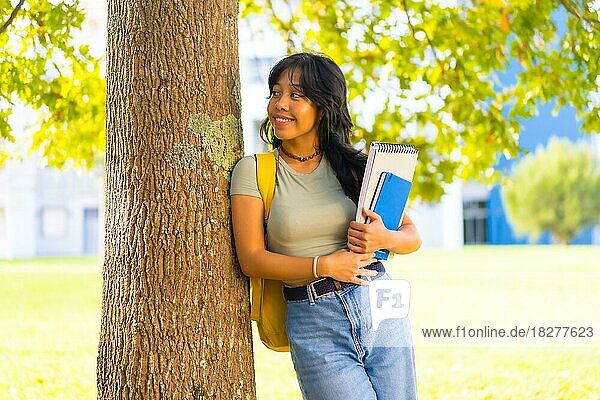 Asiatisches Mädchen auf dem Campus  lächelnd an der Universität unter einem Baum mit einem Block in der Hand