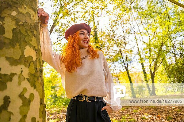 Red-haired woman leaning against a tree in a forest park smiling at sunset