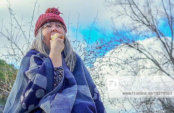 White-haired woman with red cap covered with a blanket eating an apple in the mountains