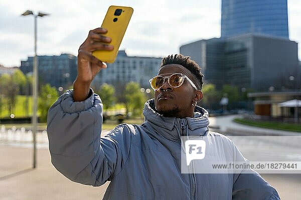 A black ethnic man with a phone and a blue jacket in the city  taking a selfie