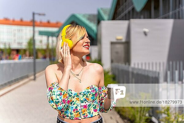 Eine hübsche blonde Frau  die mit gelben Kopfhörern und einem Kaffee zum Mitnehmen in der Stadt Musik hört  Lebensstil eines Models in der Stadt