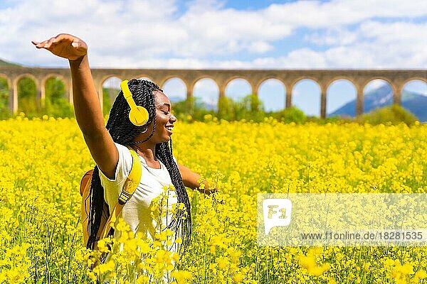 Ein Mädchen schwarzer Herkunft mit Zöpfen  eine Reisende  tanzt zu Musik mit gelben Kopfhörern und geschlossenen Augen in einem gelben Blumenfeld