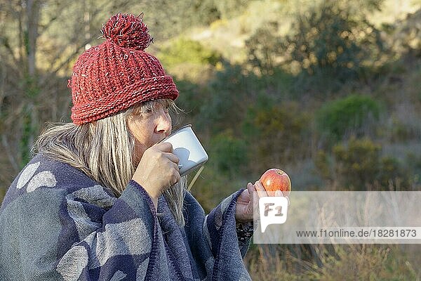 Frau mit weißen Haaren und roter Mütze trinkt Kaffee und isst einen Apfel auf einem Feld mit Bäumen im Hintergrund