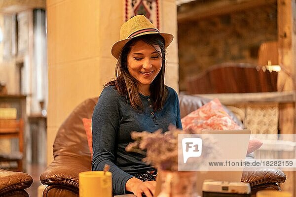 Eine hübsche Touristin mit Hut und Fotoapparat  die in einer Cafeteria Tee trinkt und im Urlaub mit einem Computer aus der Ferne arbeitet