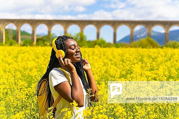 Musik hörend mit gelben Kopfhörern und geschlossenen Augen  ein schwarzes ethnisches Mädchen mit Zöpfen  eine Reisende  in einem Feld mit gelben Blumen