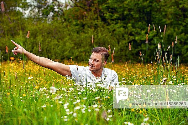 Mann sitzt in einem Blumenfeld mit ausgestreckten Arm  nach rechts zeigend