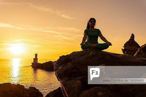 Eine Frau macht Meditations- und Yogaübungen bei Sonnenuntergang neben einem Leuchtturm im Meer  gesundes und naturistisches Leben  Pilates im Freien  ardha padmasana