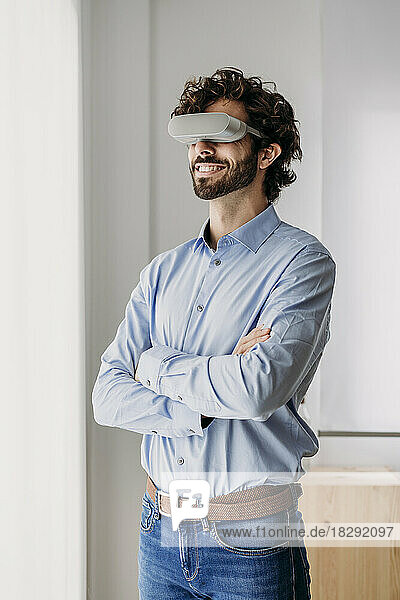 Smiling businessman wearing VR glasses standing with arms crossed in office
