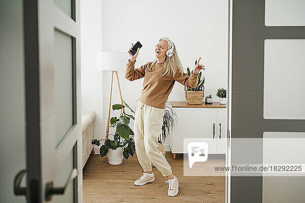 Fröhliche reife Frau mit kabellosen Kopfhörern tanzt zu Hause