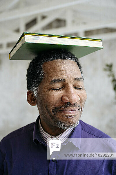 Smiling mature man with eyes closed balancing book on head