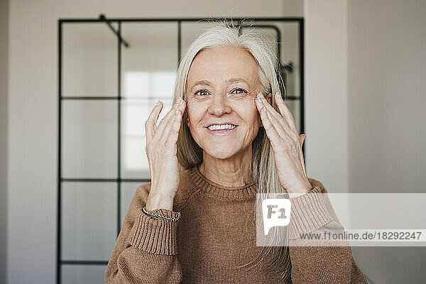 Happy mature woman applying moisturizer on face at home
