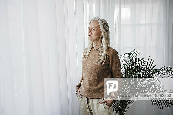 Thoughtful mature woman with hands in pockets standing by window at home