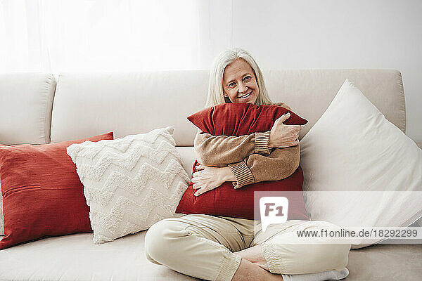 Happy mature woman with cushion sitting on sofa at home