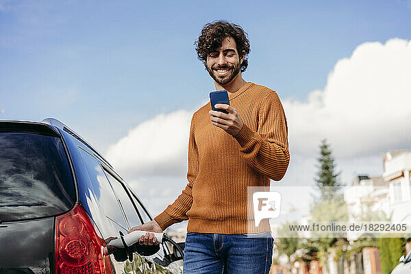 Glücklicher Mann benutzt Smartphone und lädt Auto an Fahrzeugladestation