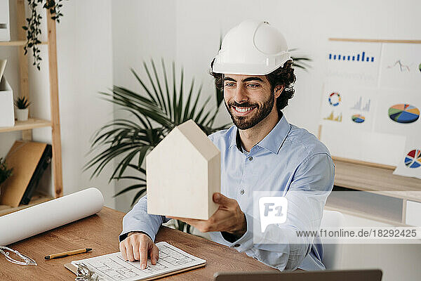 Smiling architect with blueprint holding house model at desk in office