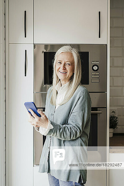 Glückliche reife Frau mit Smartphone  die in der Küche steht